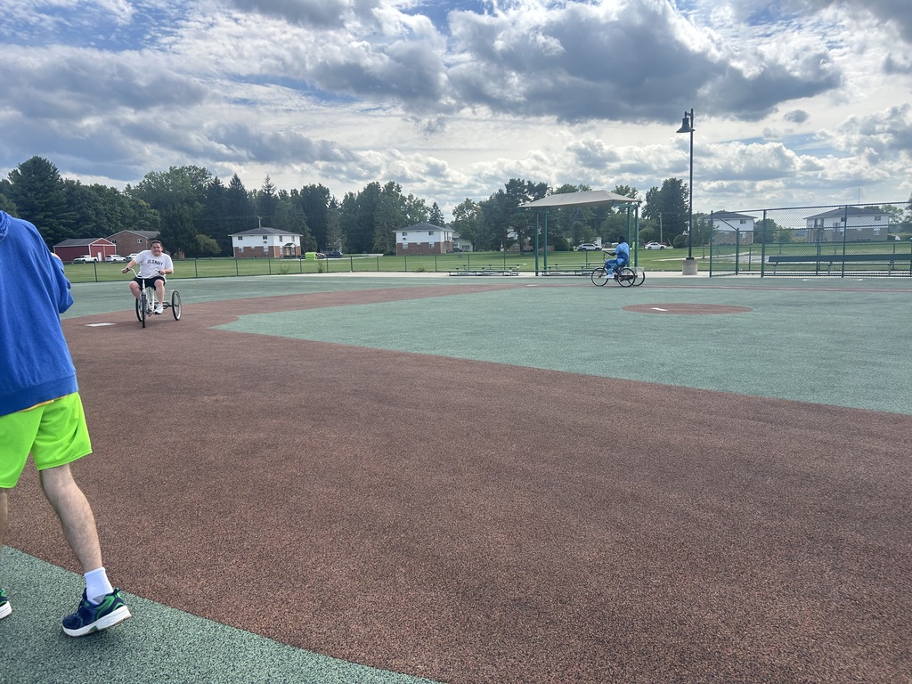 A person rides a bicycle on an outdoor sports field. The field has a mix of brown and green surfaces.