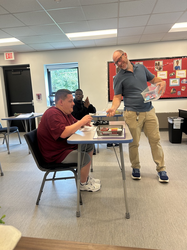 Two men in a room with desks. One man is standing at a table holding a container, the other is seated.