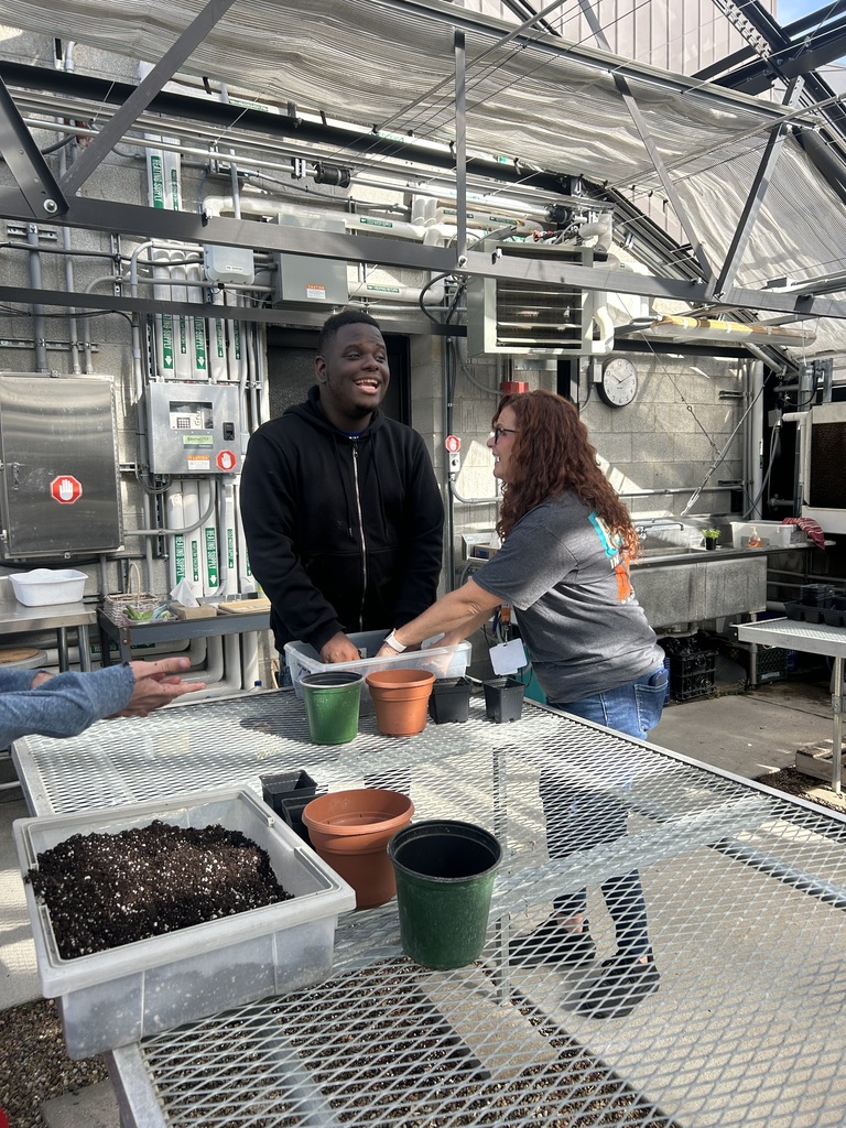 A man and a woman in a greenhouse. The man smiles. A woman holds a container.