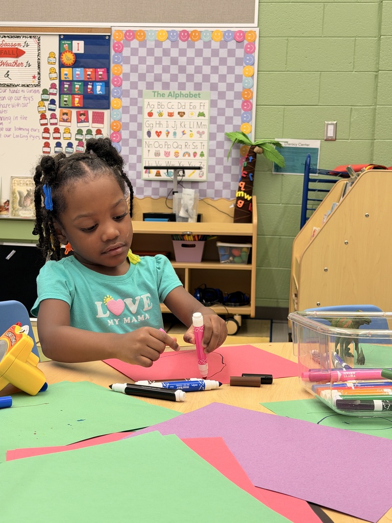 A young child sits at a table, surrounded by colored paper and art supplies. Various colored papers are on the table. Behind the child, a wall with colorful patterns and posters is visible.