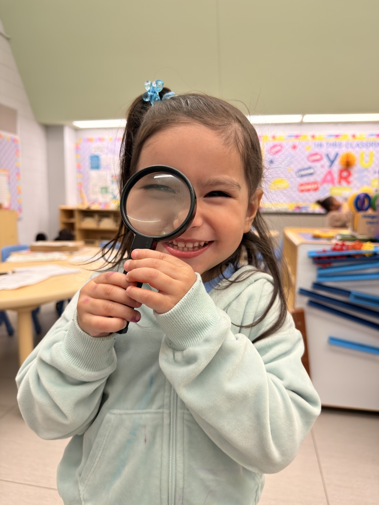 A girl with a ponytail holds a magnifying glass and smiles, standing in a classroom with desks and a bulletin board.
