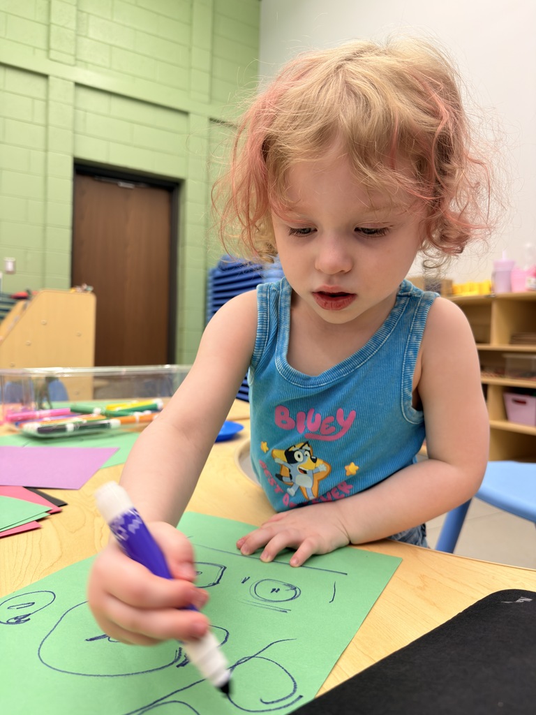 A young child with blonde hair draws on green paper with a marker, sitting at a table.