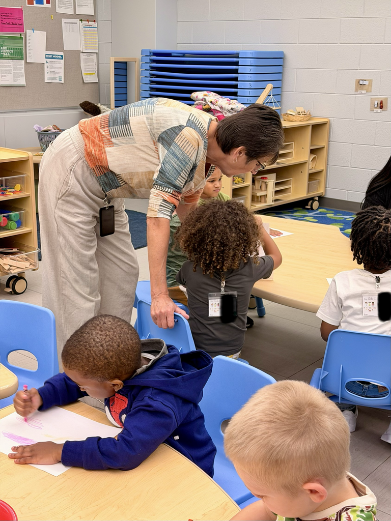 A man helps children at desks in a classroom. Some kids draw on paper. Various chairs and tables are present.