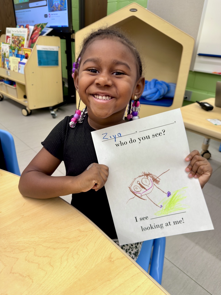 A child holds a sheet of paper with a drawing and text, "I see looking at me!" They sit in a classroom, smiling at the camera.