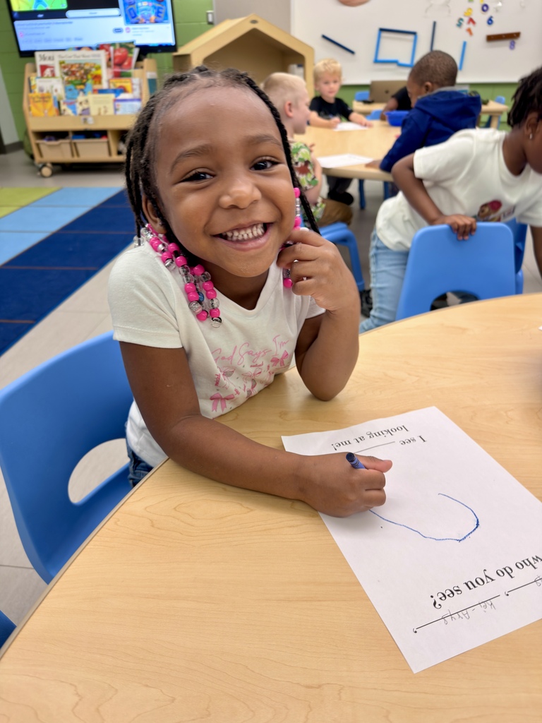 A child in a classroom sits at a table with a piece of paper, smiling. Behind her, others are seated at tables.