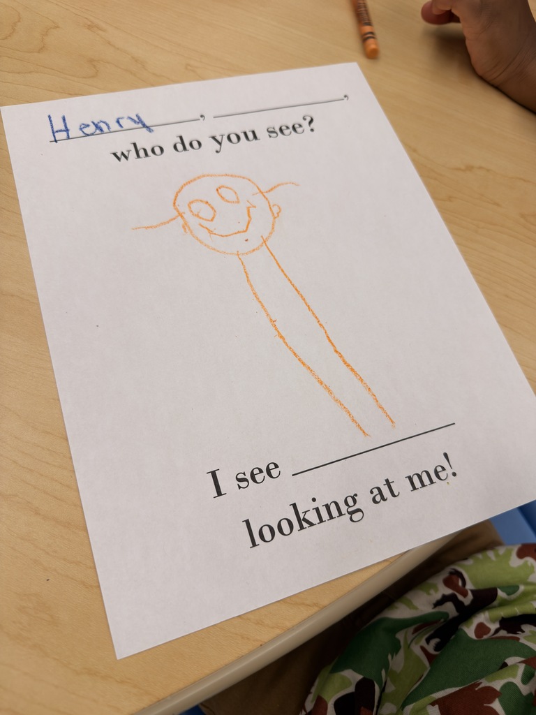A child's drawing of a smiling face on a white paper with the text "I see looking at me!" sits on a wooden table.