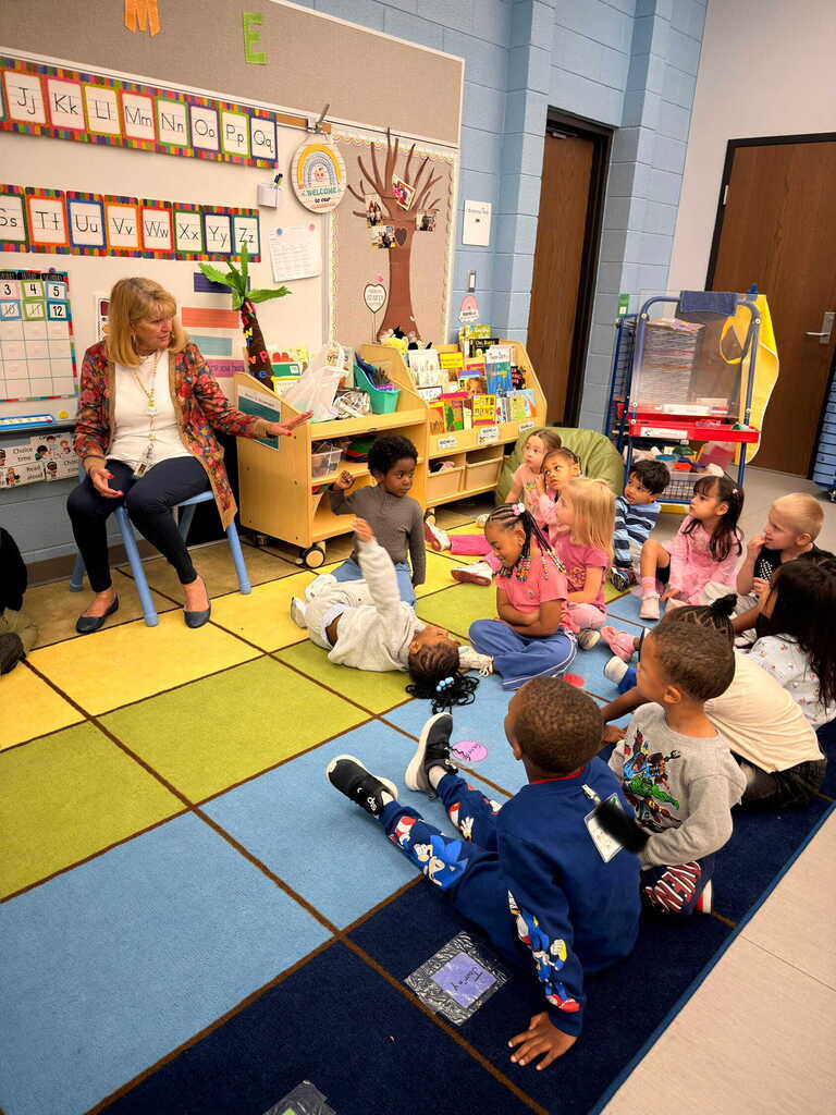 Teacher reads to students sitting in a circle on a blue mat, surrounded by colorful blocks.