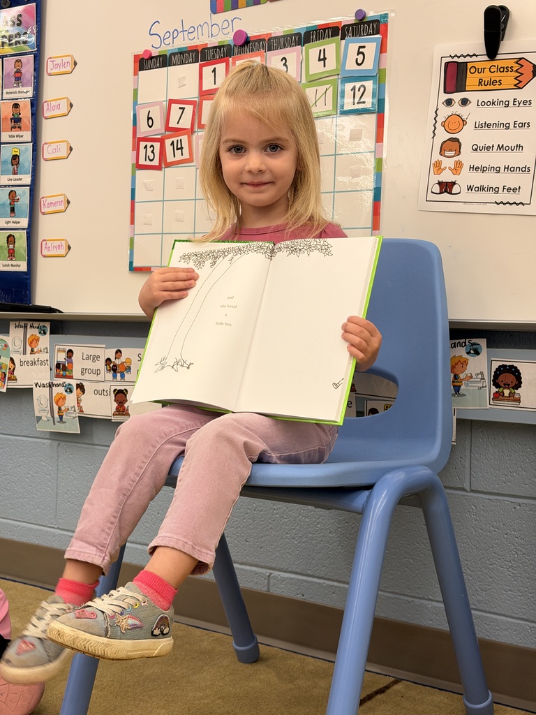 A young girl sits in a chair holding an open book, in front of a classroom calendar.