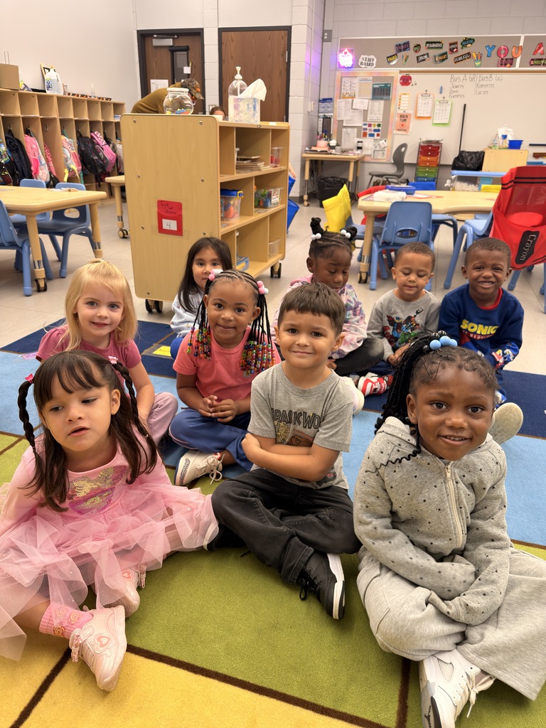 A group of children are seated on a rug inside a classroom with tables and chairs.