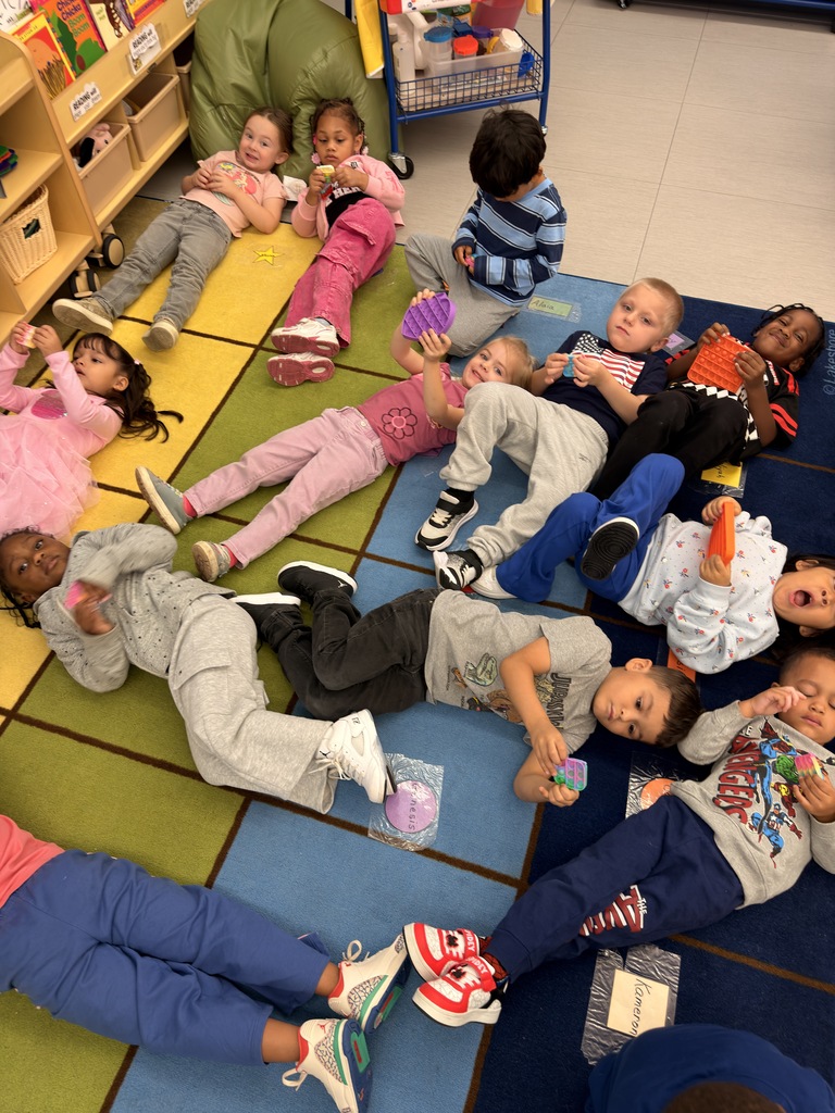 Many children lie on a colorful floor mat, some wearing sneakers and others in casual clothes.