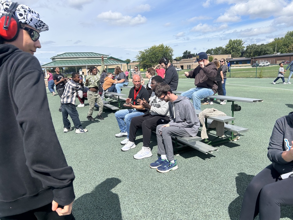 A group of people sit on benches, some in sneakers, one in headphones, with others standing on a sports field.