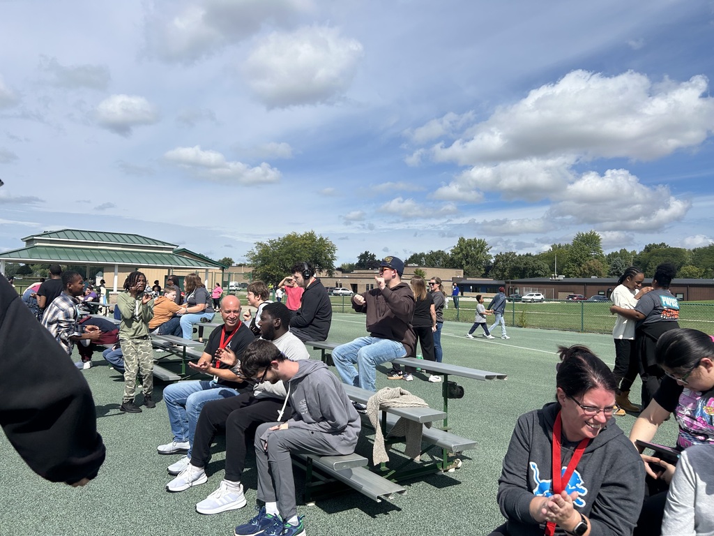 People seated on benches on a sports field under a partly cloudy sky. Some have backpacks.