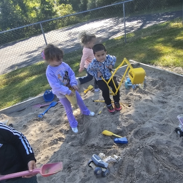 Several children play in a sandbox with toys, including a shovel and a yellow excavator.