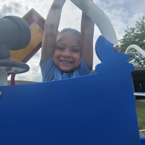 A child smiles widely while gripping the blue structure of a playground, with a tree and cloudy sky in the background.
