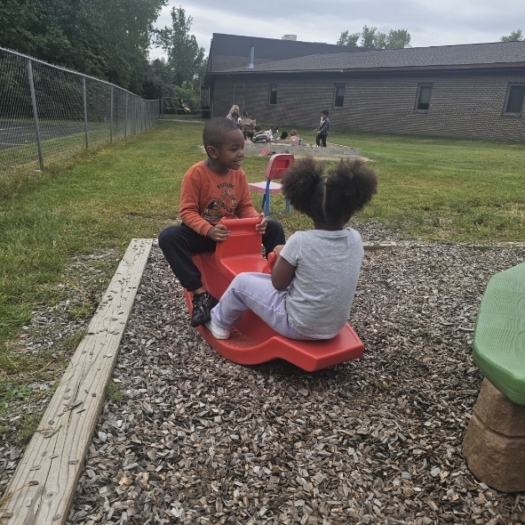 Two individuals in orange and white shirts, sitting on red plastic rides on gravel ground, with trees and a building in the background.