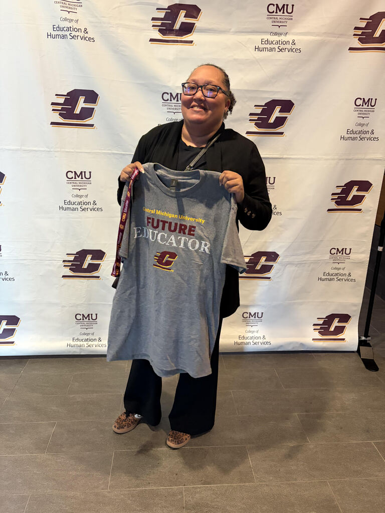 A smiling woman holds a gray t-shirt that says "FUTURE EDUCATOR" in front of a wall with logos.