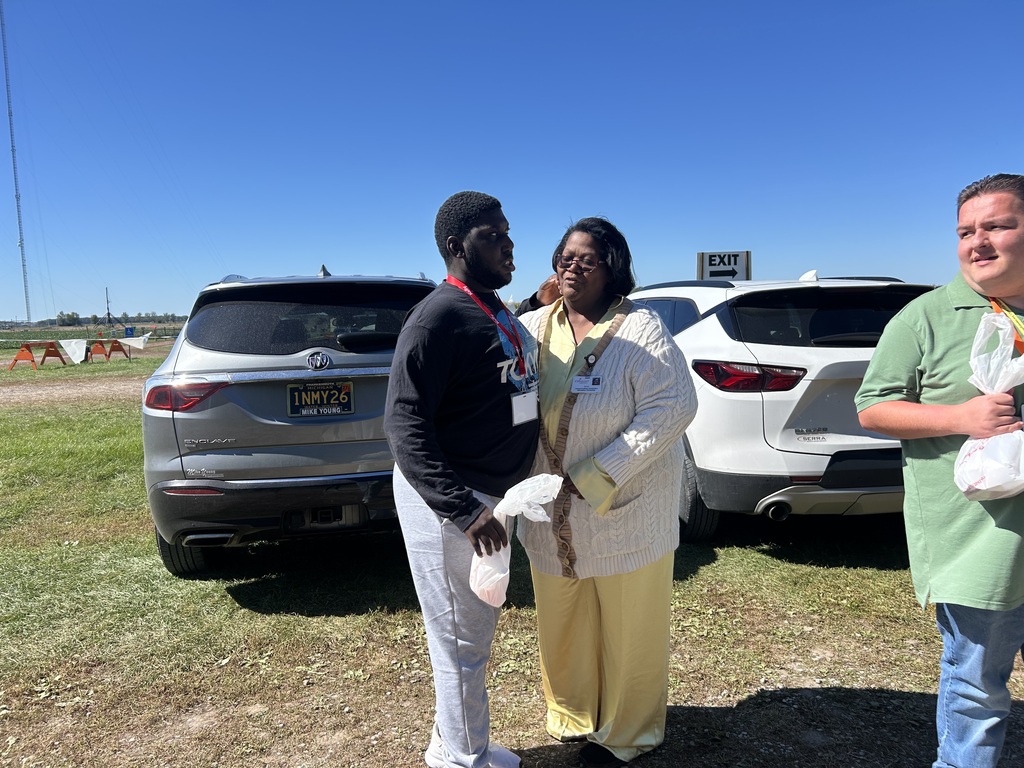 Three individuals stand in a field, two holding plastic bags, near two SUVs. Clear blue sky.