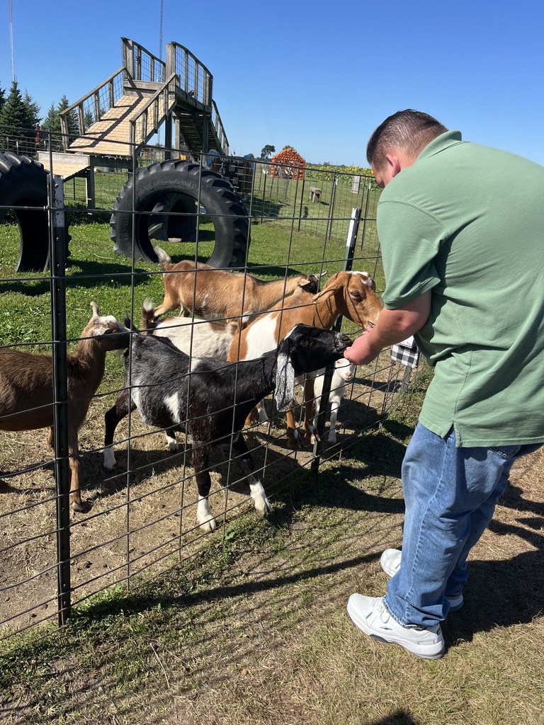 A man in a green shirt and blue jeans pets a black goat in an outdoor pen.