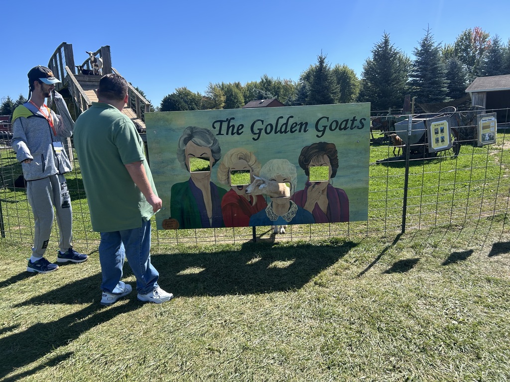 Two people stand in front of a large sign featuring four women. The sign reads "The Golden Goats." Trees and a building are in the background.