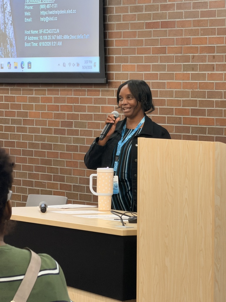 Person speaks at a podium with a microphone. A screen behind them displays text and images. Another person listens.