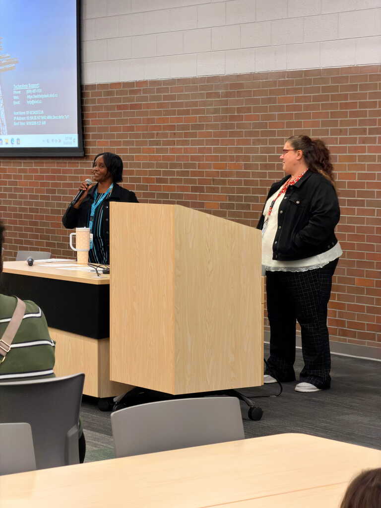 A woman speaks into a microphone at a podium. Another woman stands behind her. A projector screen displays text behind them.
