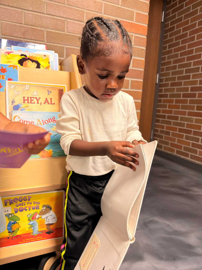 A young child in a library, holding a book, with shelves of books behind them and a brick wall.
