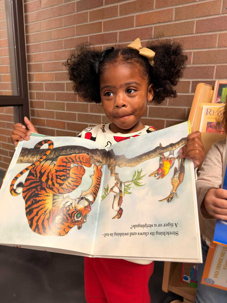 A child holding a book featuring a tiger on a branch. Brick wall and bookshelves in the background.