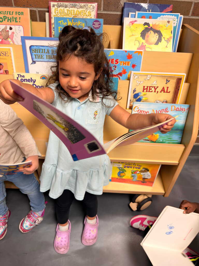 A young girl reads a book in a library. Behind her, a bookshelf filled with colorful books. Another child reads nearby.