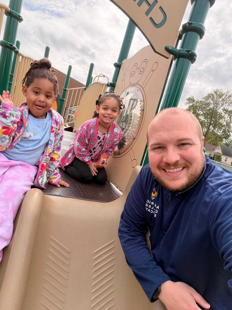 Two young girls on a playground slide with a smiling man. The background shows a cloudy sky.