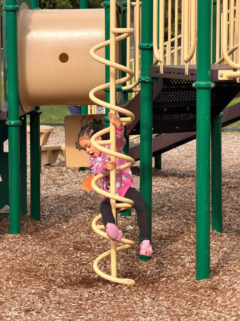 A young girl climbs a spiral yellow ladder at a playground with a slide and benches.