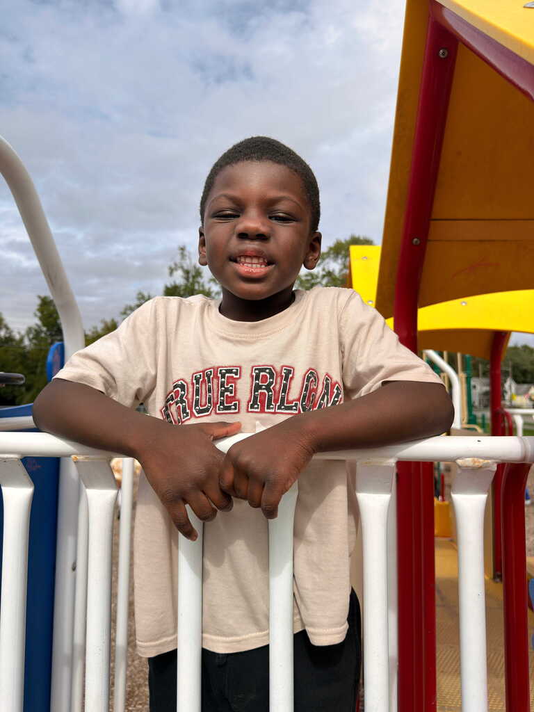 A child in a tan shirt with text on the front stands in a playground, hands on the white railing.