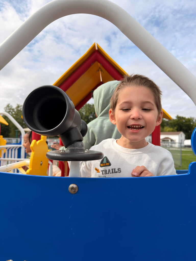 A child smiles and plays with a telescope at a playground, wearing a white shirt with "TRAILS" printed.