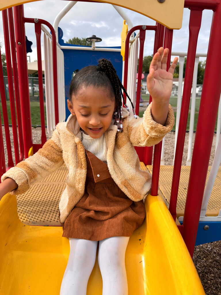 A child in a brown dress slides down a yellow slide at a playground. Bright colors and metal rails are visible.