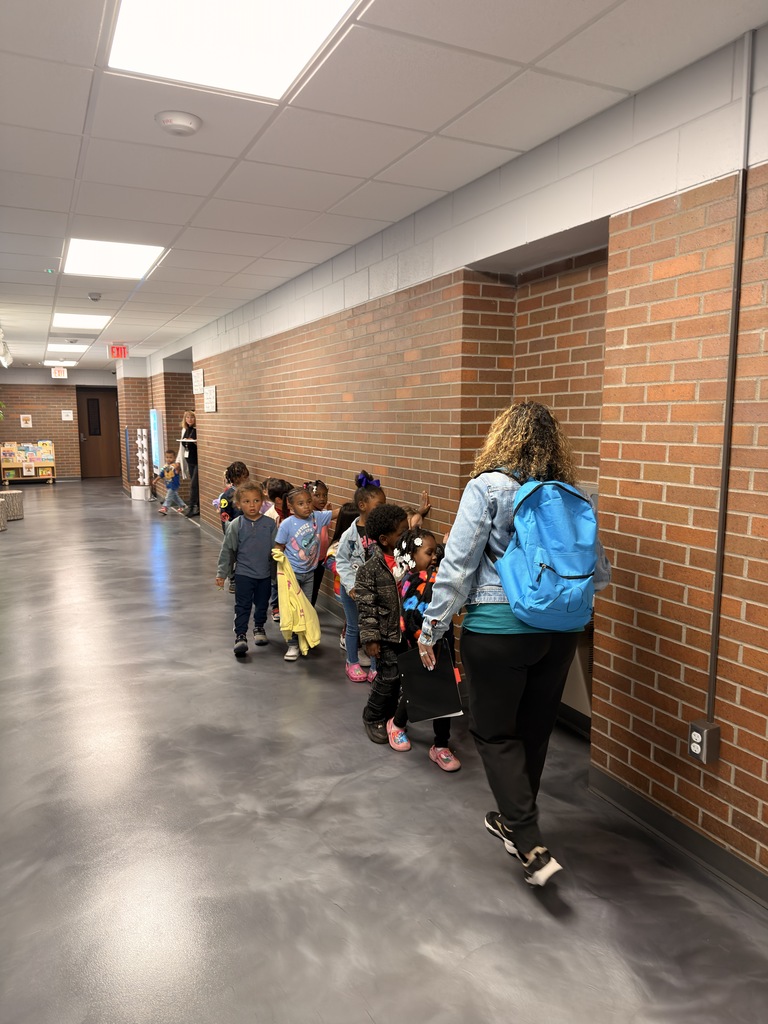 An adult and several children walk through a hallway with brick walls and fluorescent lighting.