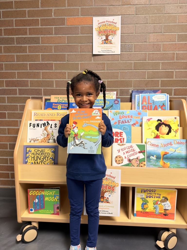 Child holding a book stands in front of a wooden bookshelf filled with children's books.