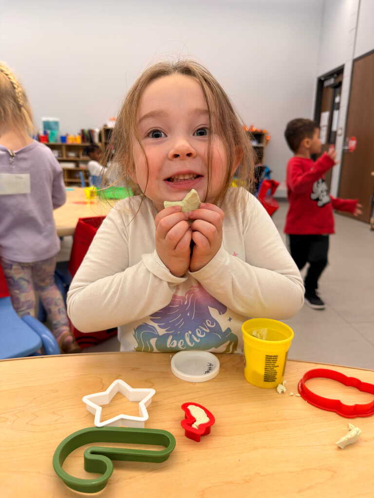 A child, seated at a table, smiles while holding a cookie cutter. Behind, a child walks, and others are present in a room with tables and chairs.