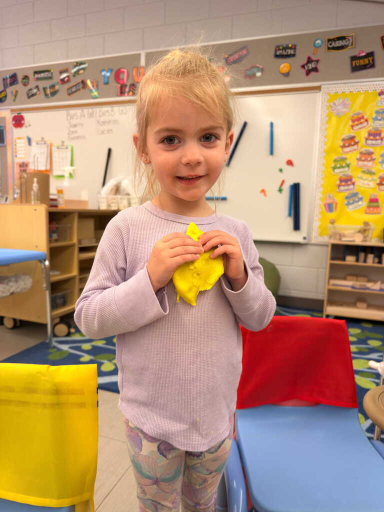 A young girl with blonde hair smiles, holding a yellow object. She stands in a classroom with chairs, shelves, and a whiteboard in the background.
