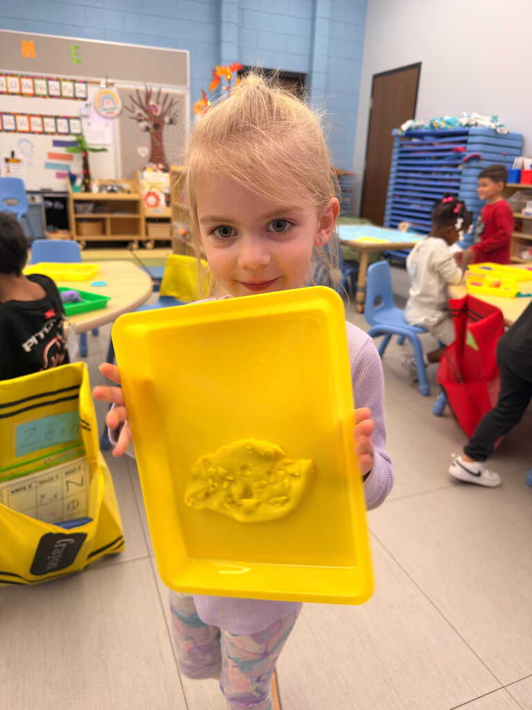 A young child holds a yellow tray with a clay figure. Behind them, a classroom setting includes tables and chairs.