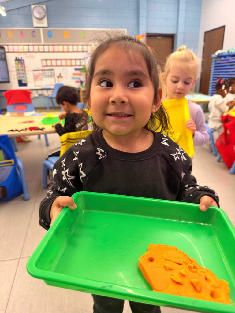 A child holds a green tray with a cookie shape on it, standing in a classroom with other kids.