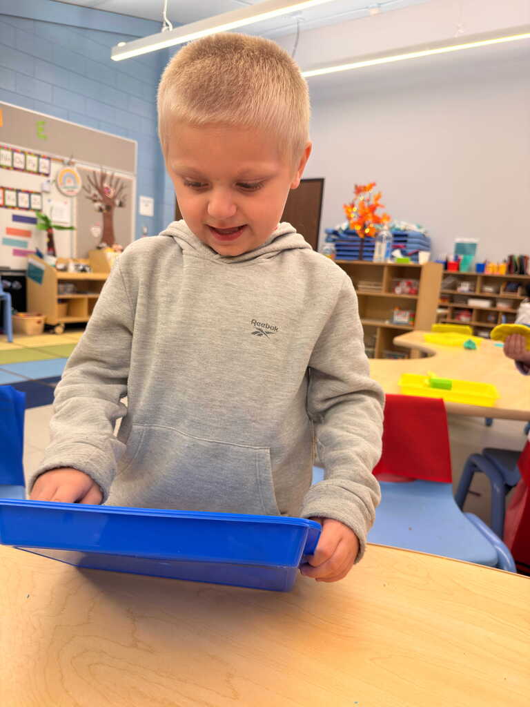 A young boy in a light gray hoodie is standing at a desk, holding a blue tray with a smile. Behind him is a classroom setting with shelves and tables.