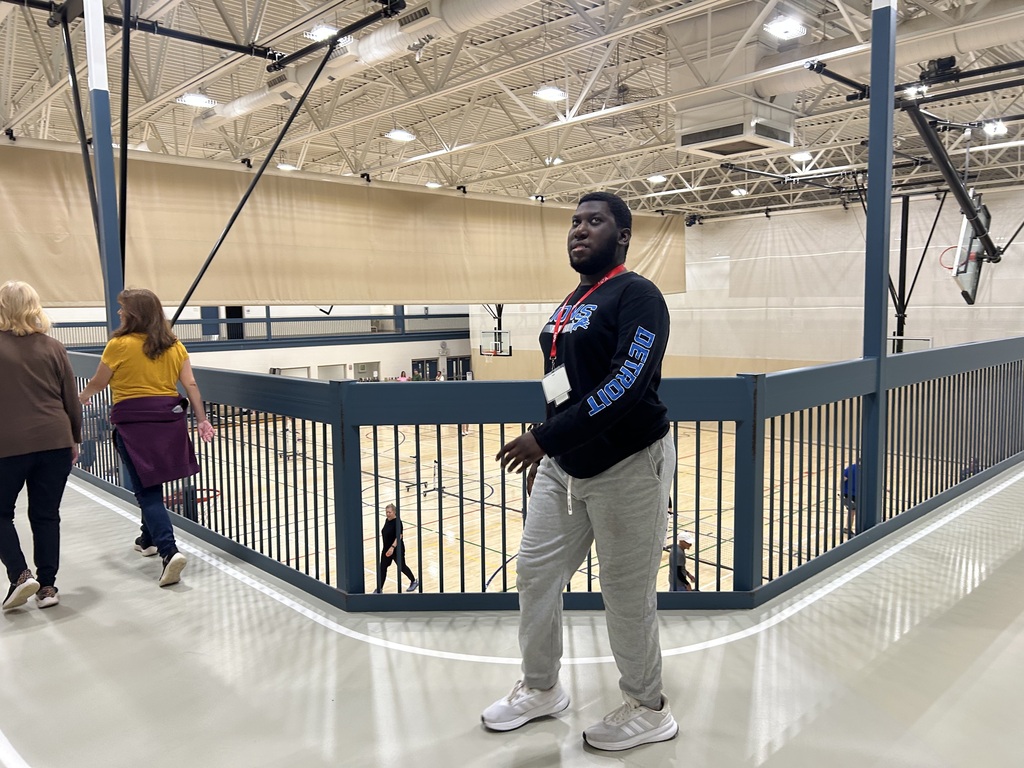 Indoor basketball court with a metal railing, a person in a sweatshirt and sneakers, and two others in front.