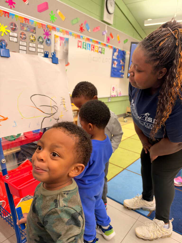 A woman helps three young children with their artwork at an easel in a classroom.