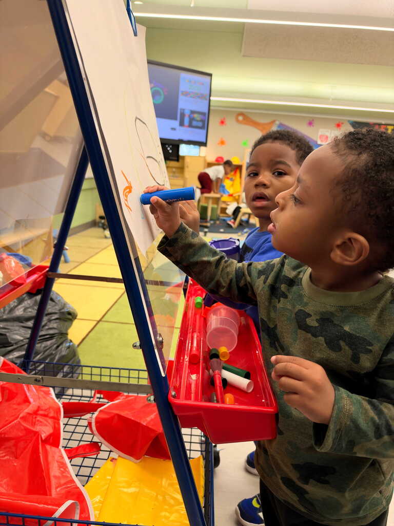 Two young children drawing on a white easel in a classroom. One child holds a blue marker.