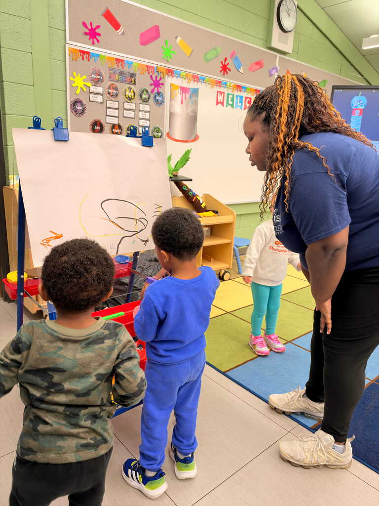 A woman watches two young boys drawing at a board in a classroom. Brightly colored walls and rugs.