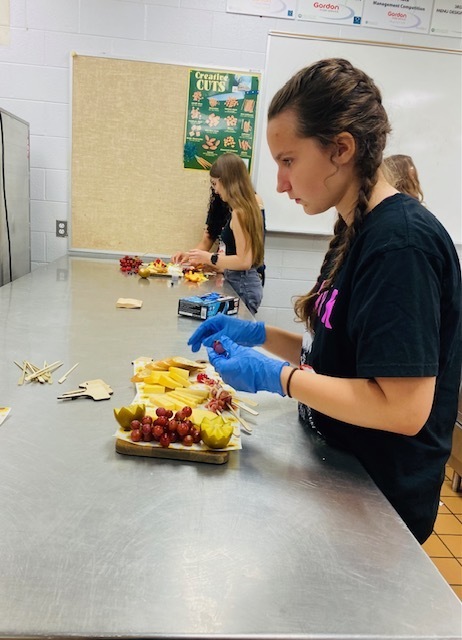 female student putting grapes on a cheese board with two students in the background