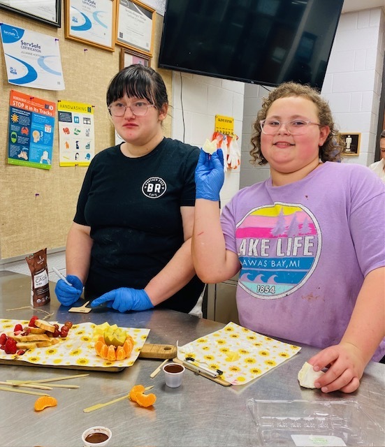 Two female students cutting up chesse and one is holding a chunk of cheese