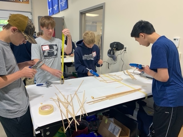Four students working at a table building a tower structure out of sticks using tape measures and hot glue guns