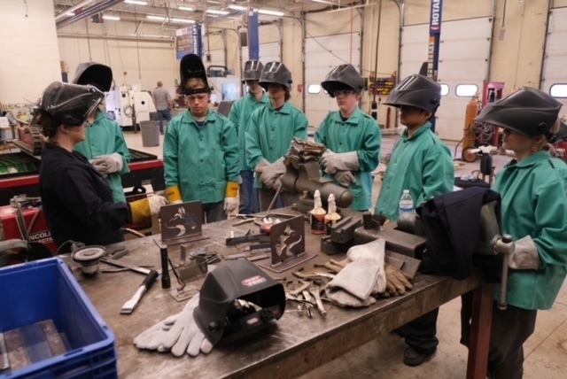 Group of 7 welding students and an instructor all dressed in welding jackets and welding helmets 