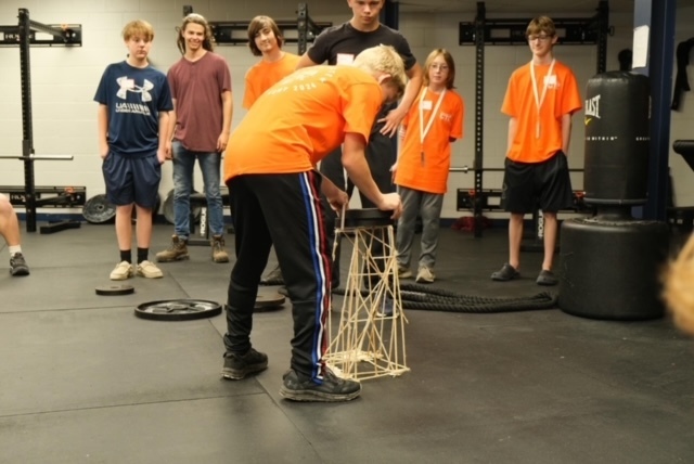 One student testing their tower structure with a 5 lb weight while other students look on to see if the tower will hold up under the pressure of the weight
