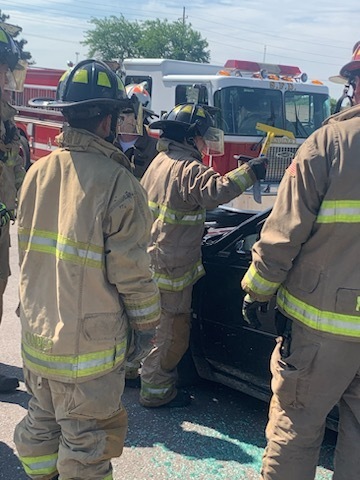 Backs of students in full fire fighter gear breaking a window on a car